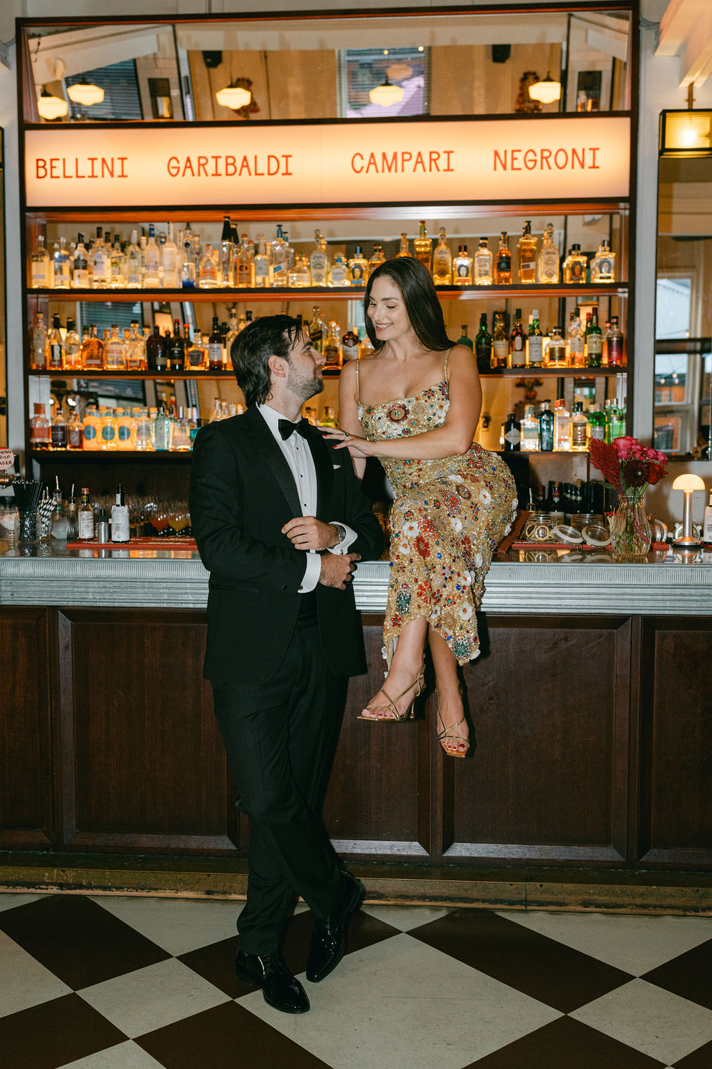 A couple in formal attire shares a moment at a bar. The woman, in a floral dress, sits on the counter, smiling warmly at the man in a tuxedo. Bottles line the shelves behind them, under a glowing sign featuring cocktail names. The setting is elegant and intimate.