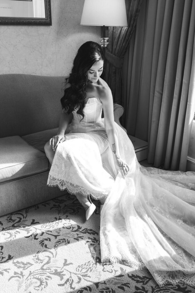 A bride in an elegant lace gown sits on a couch, illuminated by soft light. She gazes down, holding her train, creating a serene, contemplative mood.