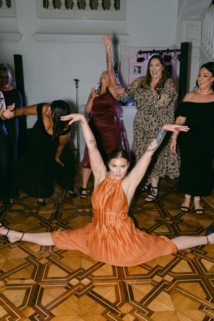A woman in an orange dress performs a split on a patterned floor, arms raised. Behind her, people cheer and dance, creating a lively party atmosphere.