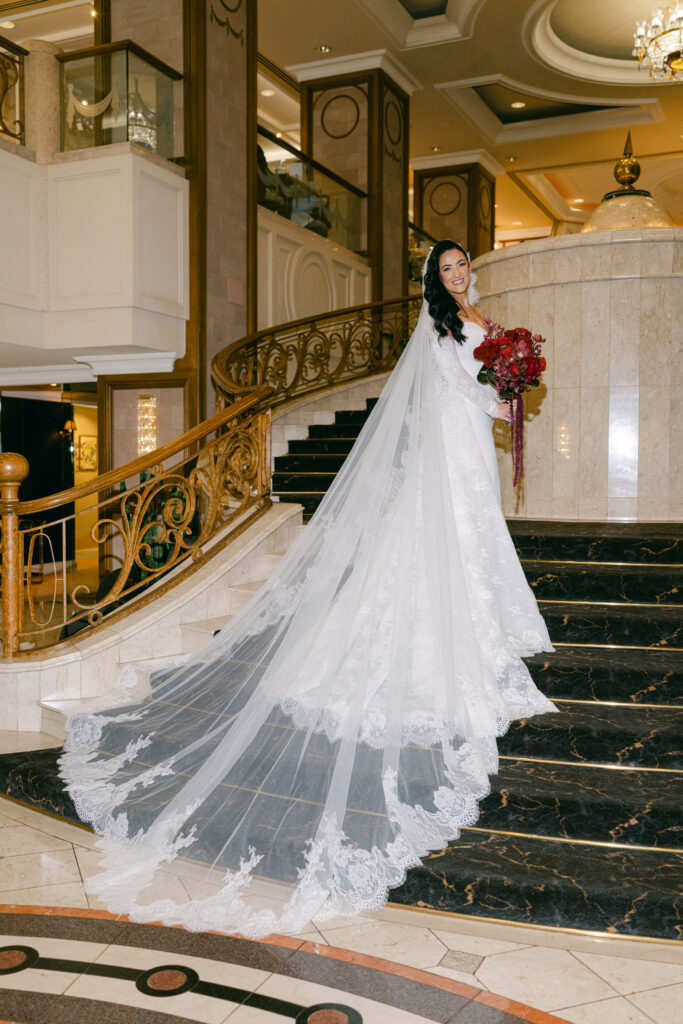 Bride in a flowing white lace gown stands on a grand staircase, holding a bouquet of red roses. The setting is elegant and opulent.