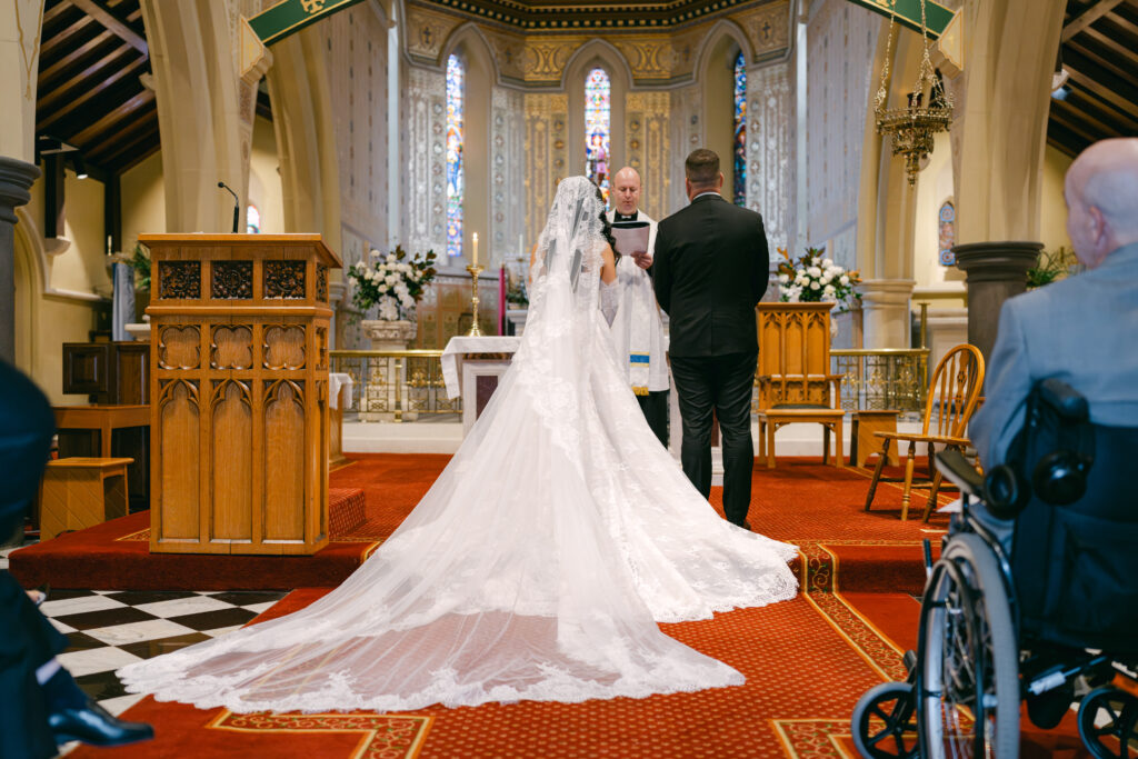 A bride and groom stand before a priest in a church with ornate stained glass and high arches. The bride's lace train flows elegantly behind her.