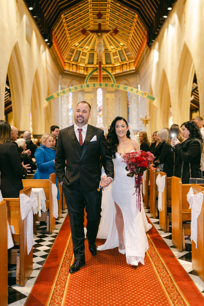 A couple walks hand in hand down a church aisle, smiling radiantly. The bride holds a vibrant red bouquet. Guests watch and smile. Joyful, elegant setting.