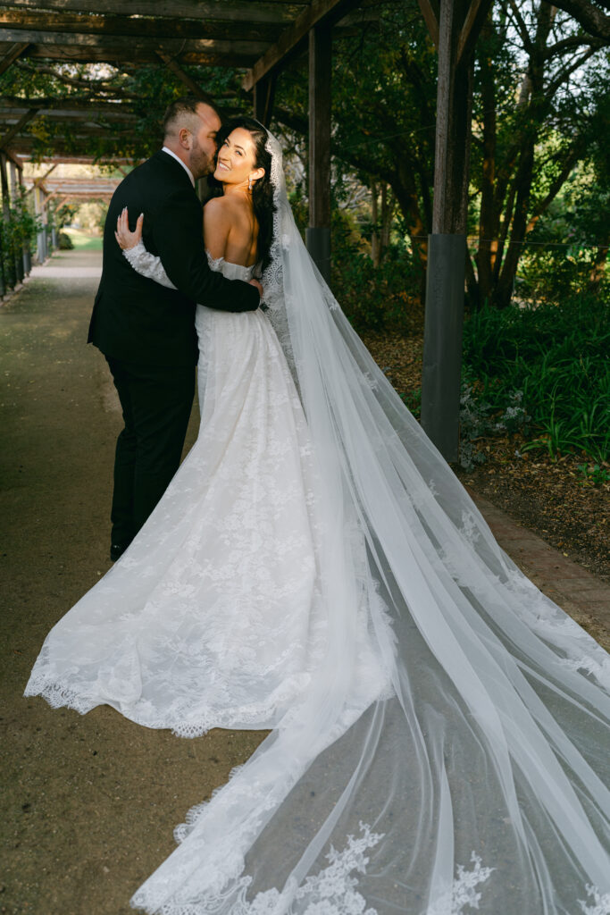 Bride and groom embrace under a wooden pergola. She wears a flowing lace gown, and he is in a black suit. The scene is romantic and serene.
