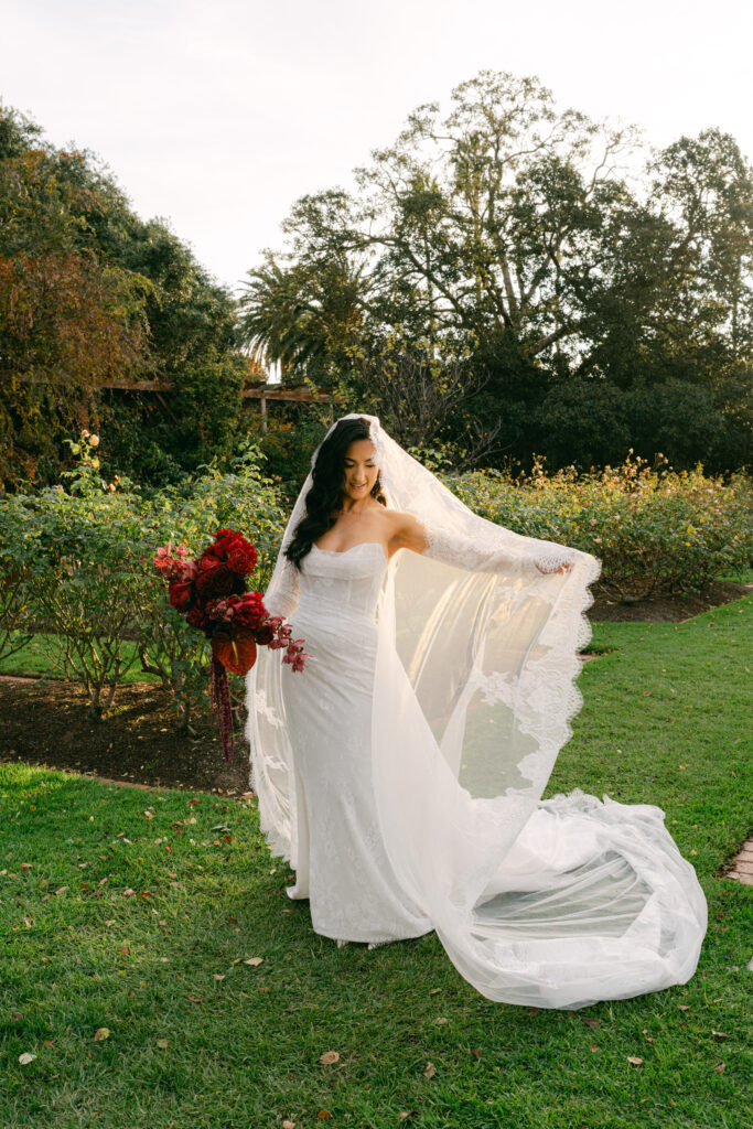 A bride in a flowing white gown and lace veil holds vibrant red flowers, standing gracefully on a green lawn amidst lush trees, exuding elegance and joy.