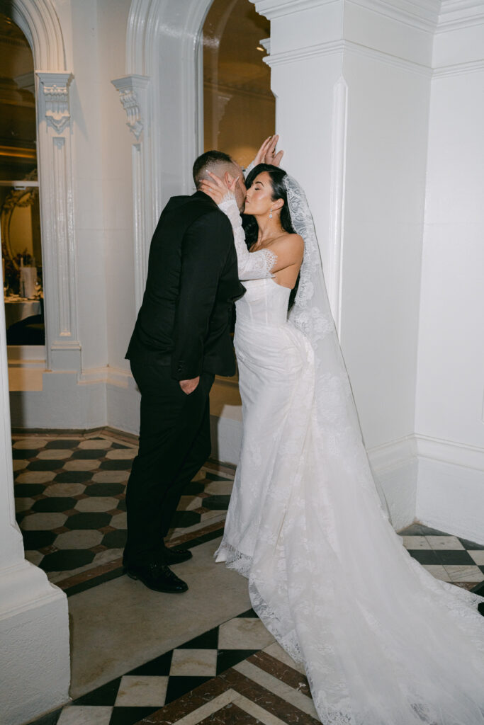 A bride and groom share a romantic kiss against a white wall. The bride wears a lace gown and veil, conveying elegance. The scene feels intimate and joyful.
