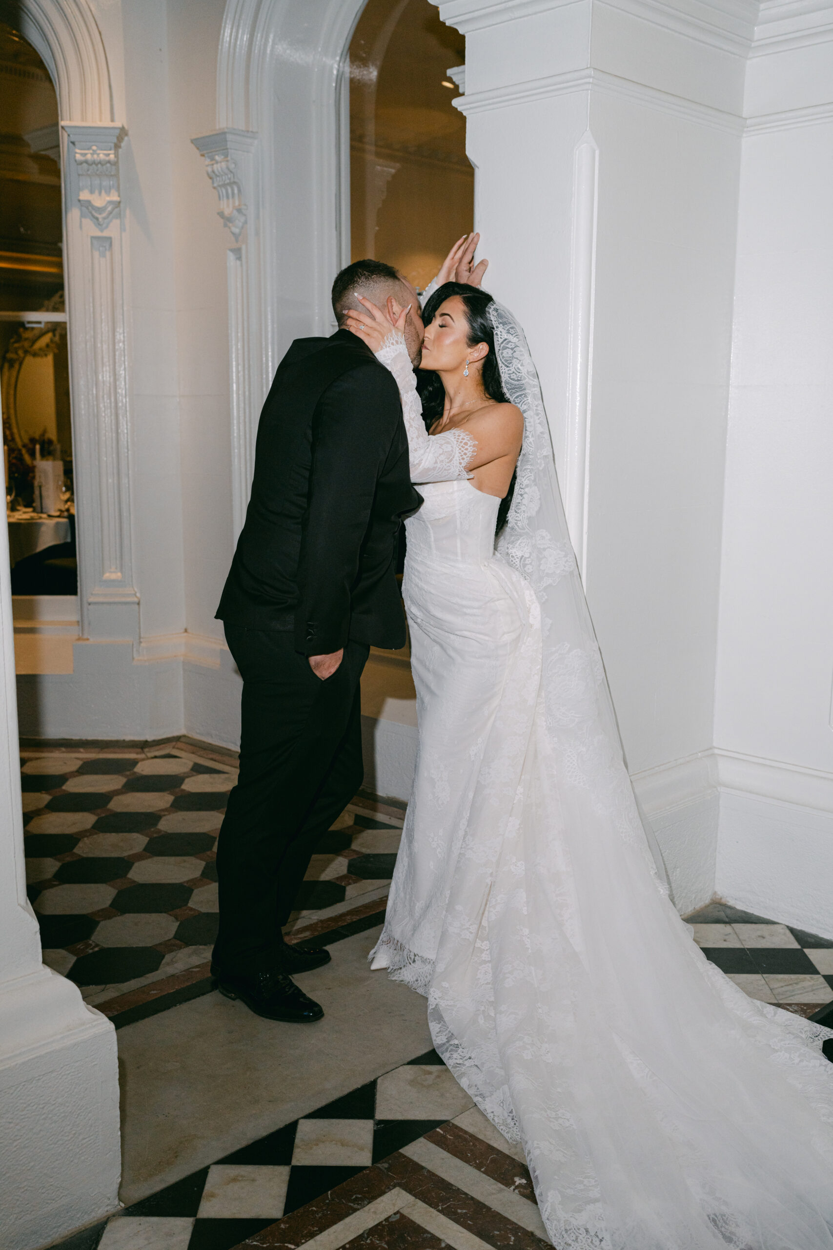 A bride and groom share a romantic kiss against a white wall. The bride wears a lace gown and veil, conveying elegance. The scene feels intimate and joyful.